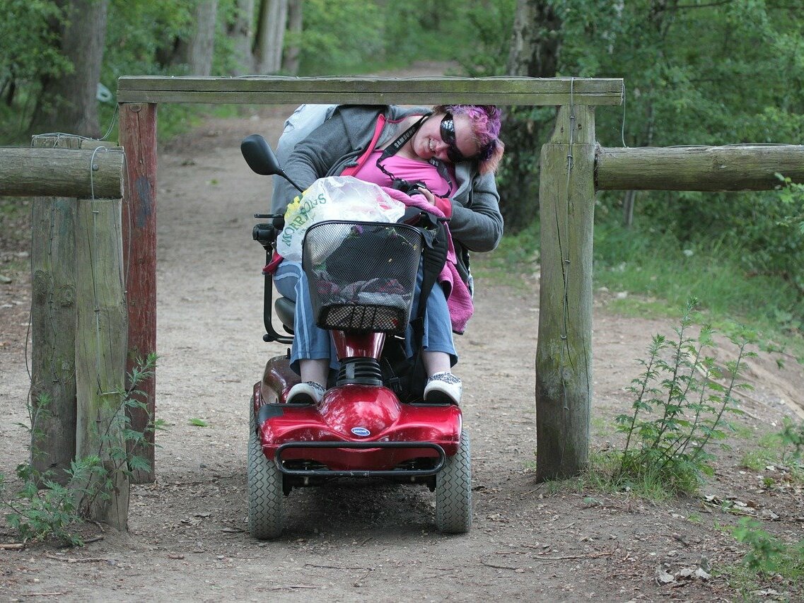 Eine Frau in einem elektrischen Rollstuhl fährt über einen Weg, an dem sie nur sehr schwer weiterkommt durch eine Verengung. 